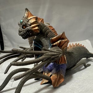 May include: A detailed figurine of a sea monster with a reptilian head, featuring a textured, scaly body in shades of gray, brown, and copper. The creature has multiple tentacles and a spiky dorsal fin, with a small, teal-colored gem on its head.