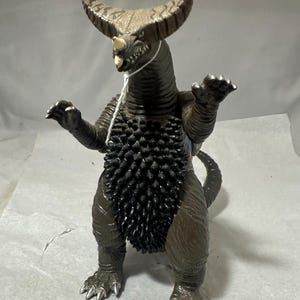 May include: A brown and black monster figurine with large horns, a spiky chest, and clawed feet. The toy has a menacing expression and is posed with its arms outstretched. The figure is a collectible item.