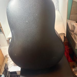 May include: A black acoustic guitar with a rounded back. The guitar's body has a textured surface. The neck of the guitar is visible at the top, with a reddish-brown finish. The guitar is resting on a dark surface.