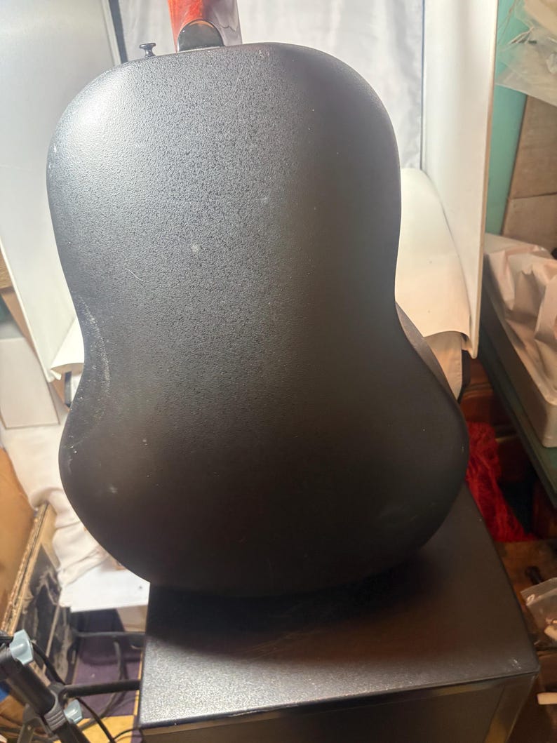 May include: A black acoustic guitar with a rounded back. The guitar's body has a textured surface. The neck of the guitar is visible at the top, with a reddish-brown finish. The guitar is resting on a dark surface.