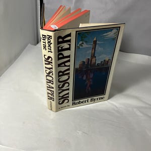 May include: A hardcover book titled "Skyscraper" by Robert Byrne. The book has a cream-colored cover with a cityscape illustration and the title in bold black letters. The pages have orange edges.