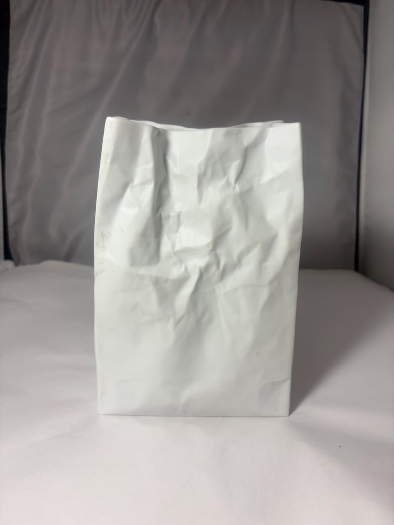 Puede incluir: Un florero de cer&aacute;mica blanca con forma de bolsa de papel arrugada. El objeto presenta un acabado mate con pliegues y arrugas marcadas en toda su superficie, imitando la apariencia de un recipiente de papel aplastado en una estructura r&iacute;gida.