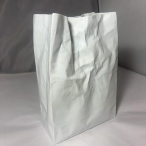 Puede incluir: Un recipiente blanco, vertical y rectangular con una superficie texturizada y arrugada que imita la apariencia de una bolsa de papel plegada. El objeto tiene una estructura r&iacute;gida con pliegues y arrugas marcadas en toda su superficie exterior.