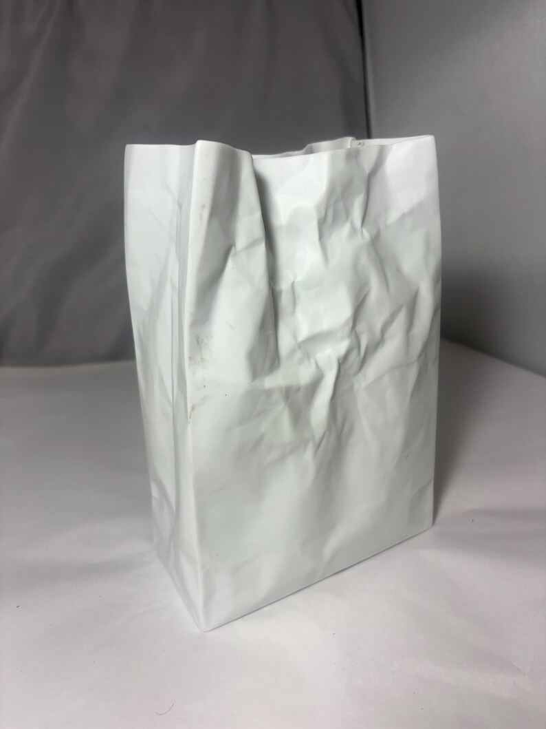 Puede incluir: Un recipiente blanco, vertical y rectangular con una superficie texturizada y arrugada que imita la apariencia de una bolsa de papel plegada. El objeto tiene una estructura r&iacute;gida con pliegues y arrugas marcadas en toda su superficie exterior.