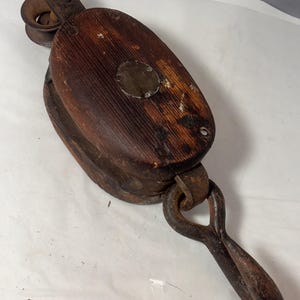 May include: Vintage wooden pulley with a dark brown finish and metal components. The pulley features a grooved wheel and a large metal loop for rope attachment. The wood shows signs of wear, giving it a rustic appearance.