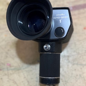 Może przedstawiać: Czarny światłomierz Minolta Auto-Spot 1° z dużą soczewką i teksturowanym uchwytem. Urządzenie posiada czarny pasek i srebrny przycisk. Widoczny jest tekst "minolta AUTO-SPOT 1°".
