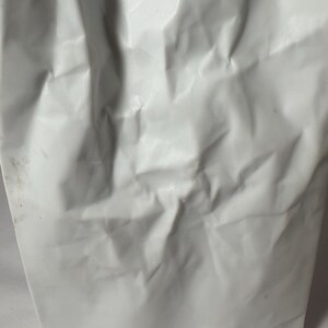 Puede incluir: Un florero blanco con una superficie texturizada que imita la apariencia de una bolsa de papel arrugada. El objeto presenta pliegues, arrugas y hendiduras irregulares en todo su exterior, creando una forma r&iacute;gida y tridimensional.