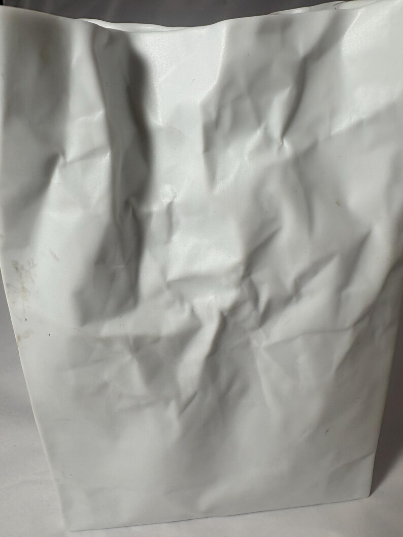 Puede incluir: Un florero blanco con una superficie texturizada que imita la apariencia de una bolsa de papel arrugada. El objeto presenta pliegues, arrugas y hendiduras irregulares en todo su exterior, creando una forma r&iacute;gida y tridimensional.