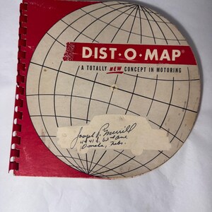 May include: A vintage spiral-bound Dist-O-Map book with a red spine and a circular off-white cover featuring a black grid globe design. The text reads Dist-O-Map A Totally New Concept In Motoring with a handwritten name and address on a paper label.
