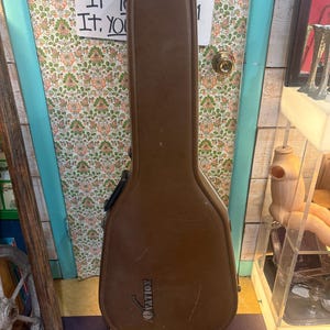 May include: A brown guitar case with the brand name "Ovation" printed on the front. The case is made of a durable material and has a handle for carrying. The case is standing in front of a floral patterned door. The text "IF YOU IT. YOU" is visible on the door.