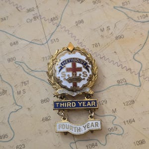 May include: A gold-tone metal pin featuring a central white emblem with a red cross, a crown, and the text METHODIST S.S. Below this, a banner reads SECOND YEAR, followed by two hanging bars labeled THIRD YEAR and FOURTH YEAR in blue and white enamel.