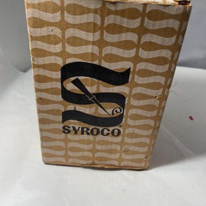 Può includere: Scatola rettangolare di cartone con un motivo a onde ripetute in beige e bianco. La scatola presenta un logo nero con la scritta "SYROCO" e un design stilizzato. La scatola è leggermente usurata.