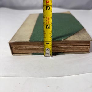 Puede incluir: Un libro antiguo con una cubierta verde y bordes color beige, que mide aproximadamente 6,35 cm de altura. Las p&aacute;ginas del libro est&aacute;n envejecidas, y una cinta m&eacute;trica amarilla se coloca junto a &eacute;l, proporcionando una referencia de tama&ntilde;o. El libro est&aacute; sobre una superficie blanca.