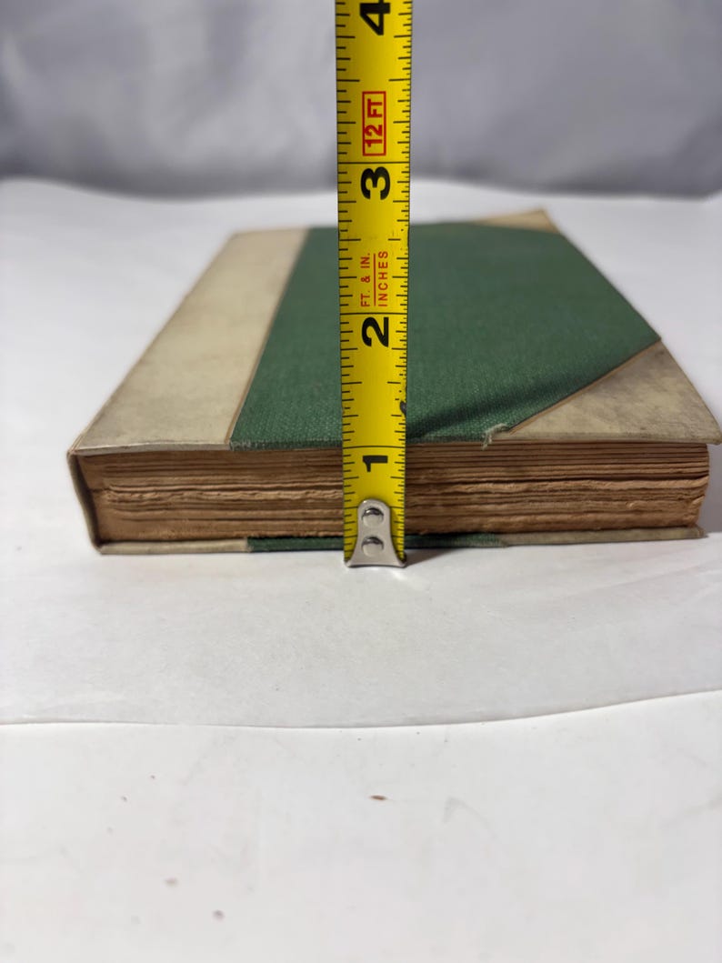 Puede incluir: Un libro antiguo con una cubierta verde y bordes color beige, que mide aproximadamente 6,35 cm de altura. Las p&aacute;ginas del libro est&aacute;n envejecidas, y una cinta m&eacute;trica amarilla se coloca junto a &eacute;l, proporcionando una referencia de tama&ntilde;o. El libro est&aacute; sobre una superficie blanca.