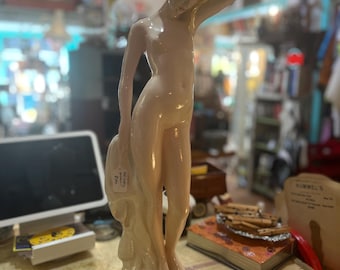 May include: A ceramic figurine of a nude woman, with her arm raised to her face. The statue is a pale, flesh-toned color with a glossy finish. The woman's hair is styled in a bun. The figurine is standing on a round, white base.