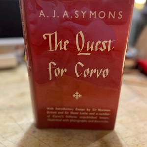 May include: A red book titled "The Quest for Corvo" by A. J. A. Symons. The title is in a decorative font, with additional text below. The book has a decorative emblem and is illustrated with photographs and documents.
