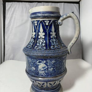 Może przedstawiać: Niebiesko-biały ceramiczny dzban z uchwytem. Dzban zdobią skomplikowane wzory kwiatowe i geometryczne, z detalicznym wzorem reliefowym wokół korpusu. Uchwyt jest łukowaty i zdobiony. Dzban ma szeroką podstawę i wąską szyję.