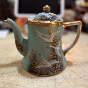 May include: A vintage, light blue porcelain teapot with a gold handle, spout, and trim. The teapot features a lid and is decorated with white swans and gold floral accents. The teapot is a decorative item.