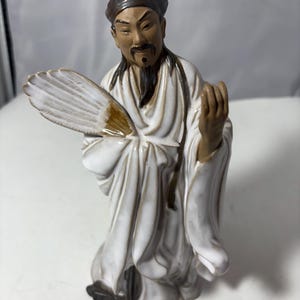 May include: A ceramic figurine of an Asian man in traditional robes, holding a fan. The figure is primarily white with brown and black accents, including the hat, facial features, and the fan's details. The figurine has a serene expression.