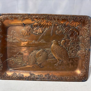 May include: A rectangular copper-toned tray with a detailed relief design. The tray features a landscape with two birds, foliage, and a water scene. The border has an ornate, repeating pattern. The overall aesthetic is vintage and decorative.