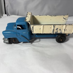 May include: A vintage metal toy dump truck featuring a blue cab and a cream-colored dump bed. The paint on the dump bed shows significant wear and chipping, revealing the metal underneath. The truck has black wheels and a silver-colored front bumper.
