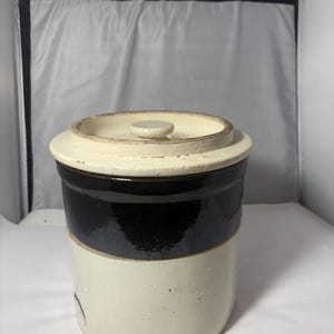 Op de afbeelding: Een vintage steengoed pot met een deksel. De pot heeft een crèmekleurige basis, een brede band van donkerbruin glazuur en een crèmekleurige rand en deksel. Het deksel heeft een kleine, ronde knop. De pot is cilindrisch en lijkt in goede staat te verkeren.