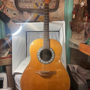 May include: Acoustic guitar with a light brown finish. The guitar has a round sound hole with a decorative rosette and a wooden bridge. The headstock is visible, and the strings are strung across the body. The guitar is in a well-lit room.