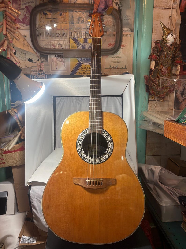 May include: Acoustic guitar with a light brown finish. The guitar has a round sound hole with a decorative rosette and a wooden bridge. The headstock is visible, and the strings are strung across the body. The guitar is in a well-lit room.