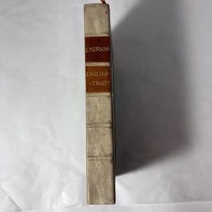 Puede incluir: Un libro antiguo titulado "English Traits" de Emerson. El libro tiene una cubierta beige claro con un lomo marr&oacute;n y naranja. El t&iacute;tulo y el nombre del autor est&aacute;n impresos en letras doradas. Una cinta roja y naranja como marcap&aacute;ginas sobresale de la parte superior.