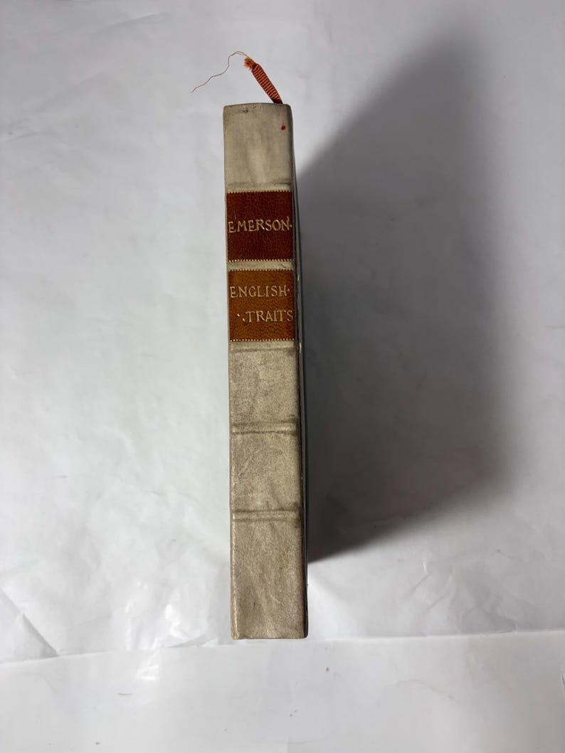 Puede incluir: Un libro antiguo titulado "English Traits" de Emerson. El libro tiene una cubierta beige claro con un lomo marr&oacute;n y naranja. El t&iacute;tulo y el nombre del autor est&aacute;n impresos en letras doradas. Una cinta roja y naranja como marcap&aacute;ginas sobresale de la parte superior.
