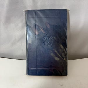 May include: A navy blue book, wrapped in clear plastic. The cover has a textured, grid-like pattern with a decorative emblem in the center. The book appears to be a hardcover edition.