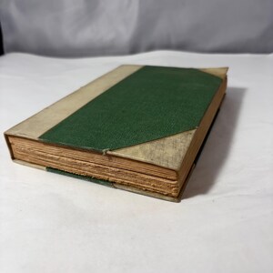 Puede incluir: Un libro antiguo con lomo verde texturizado y cubiertas beige. Las p&aacute;ginas est&aacute;n envejecidas, con un borde de pliego visible. El libro est&aacute; ligeramente abierto, revelando el papel envejecido.