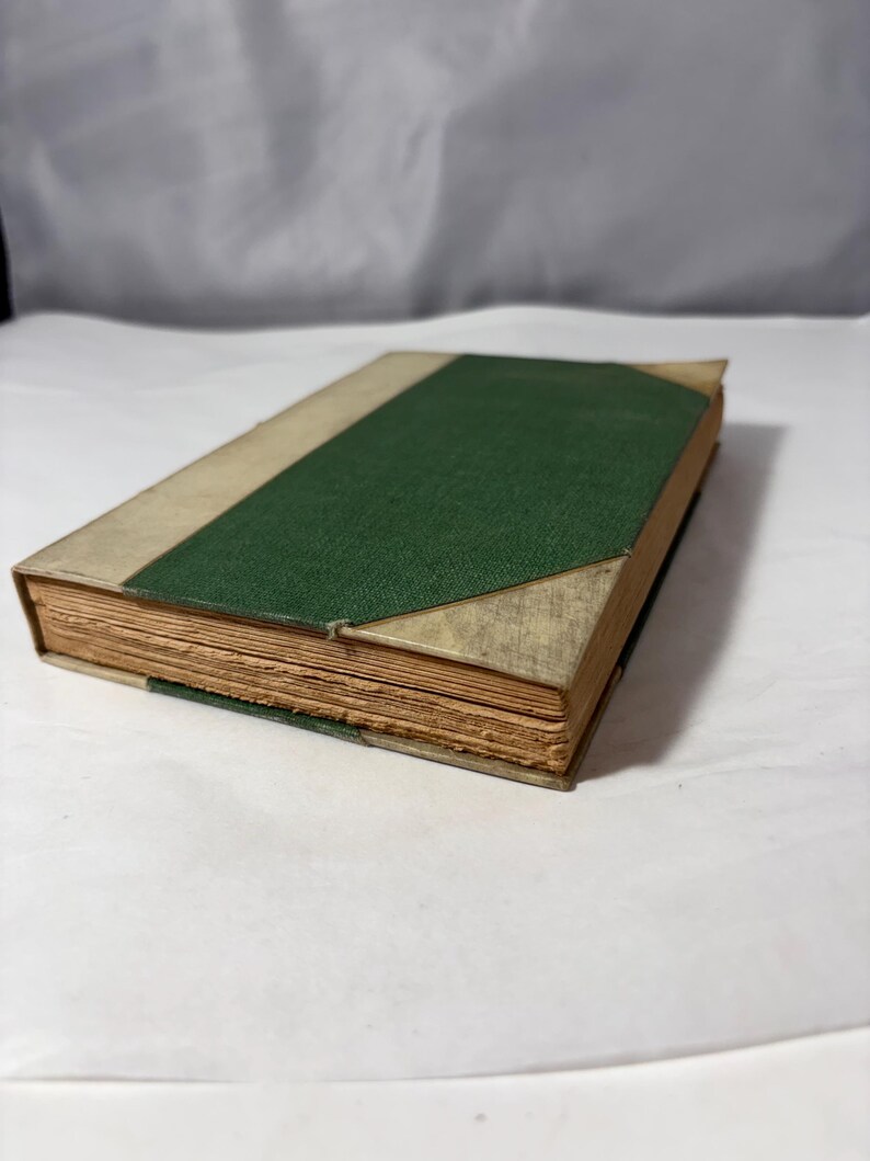 Puede incluir: Un libro antiguo con lomo verde texturizado y cubiertas beige. Las p&aacute;ginas est&aacute;n envejecidas, con un borde de pliego visible. El libro est&aacute; ligeramente abierto, revelando el papel envejecido.