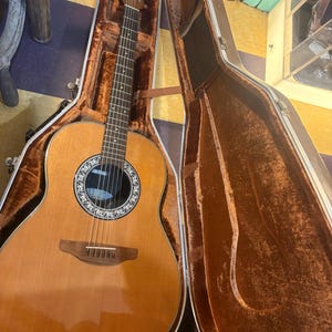 May include: Acoustic guitar in a brown case. The guitar has a light brown wooden body with a round sound hole and a black and white rosette. The case is open, revealing a brown velvet interior.