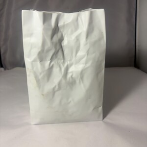 Puede incluir: Un jarr&oacute;n de cer&aacute;mica blanco con forma de bolsa y una textura arrugada similar al papel. El objeto presenta una base rectangular y una abertura superior con bordes irregulares y plegados, imitando la apariencia de una bolsa de papel arrugada.