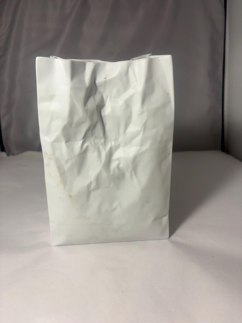 Puede incluir: Un jarr&oacute;n de cer&aacute;mica blanco con forma de bolsa y una textura arrugada similar al papel. El objeto presenta una base rectangular y una abertura superior con bordes irregulares y plegados, imitando la apariencia de una bolsa de papel arrugada.