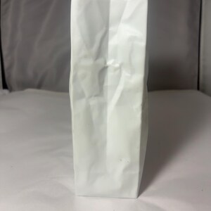 Puede incluir: Un jarr&oacute;n rectangular alto de cer&aacute;mica blanca con una superficie texturizada que imita la apariencia de una bolsa de papel plegada. El objeto presenta pliegues verticales marcados y un acabado mate, manteni&eacute;ndose erguido con una base plana.