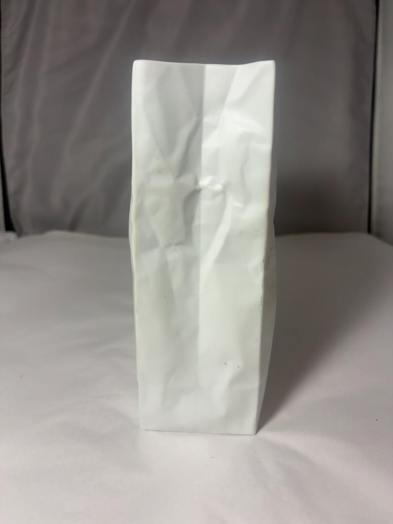 Puede incluir: Un jarr&oacute;n rectangular alto de cer&aacute;mica blanca con una superficie texturizada que imita la apariencia de una bolsa de papel plegada. El objeto presenta pliegues verticales marcados y un acabado mate, manteni&eacute;ndose erguido con una base plana.