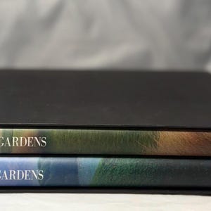 May include: Two books titled "THE WIRTZ GARDENS" are stacked inside a black box. The top book has a cover with brown and green hues, while the bottom book has blue and green tones. The books are in a black box.