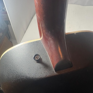 May include: Close-up of the neck and body of an acoustic guitar. The neck is a dark reddish-brown color, and the body is black with a textured surface. A single screw is visible near the neck joint.