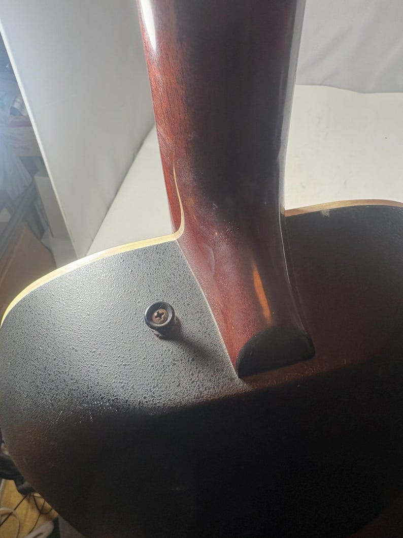 May include: Close-up of the neck and body of an acoustic guitar. The neck is a dark reddish-brown color, and the body is black with a textured surface. A single screw is visible near the neck joint.