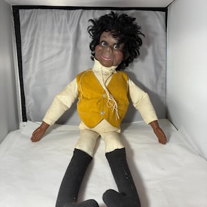 Puede incluir: Un muñeco de ventrílocuo de piel oscura, cabello negro rizado y una amplia sonrisa. El muñeco viste una camisa de manga larga color crema, un chaleco amarillo mostaza con tres botones, pantalones color crema y botas largas de tela negra.