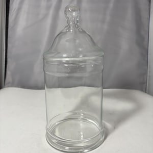 May include: A clear glass apothecary jar with a lid. The jar has a cylindrical body and a rounded base. The lid has a knob on top. The jar is sitting on a white surface.