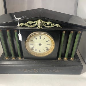 May include: A black mantel clock with a gold-rimmed clock face and Roman numerals. The clock has green and gold accents, with a decorative top and base. The clock's design is reminiscent of classical architecture.