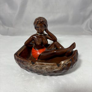 May include: A vintage ceramic ashtray featuring a reclining figure in a brown boat-shaped dish. The figure has dark brown skin, a red swimsuit, and a gold necklace. The ashtray has a brown and cream marbled design.