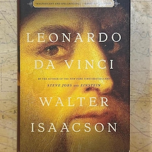 May include: A hardcover book titled Leonardo da Vinci by Walter Isaacson. The cover features a golden-toned close-up portrait of a face with the text "MAGNIFICENT AND SPELLBINDING" at the top and "BY THE AUTHOR OF THE NEW YORK TIMES BESTSELLERS STEVE JOBS AND EINSTEIN" in the center.