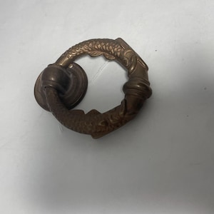 May include: Antique bronze-toned drawer pull with a circular handle. The handle features a detailed fish scale pattern and decorative accents. The pull is attached to a round backplate, showcasing a vintage design.