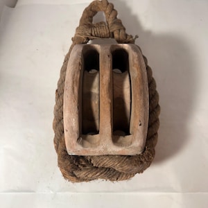 May include: A vintage wooden block and tackle pulley with a thick, woven rope. The pulley has two internal wheels and is encased in a tightly wound rope. The wood has a weathered appearance, suggesting age and use. The rope is a natural brown colour.