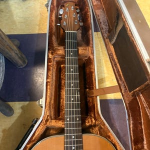 May include: An acoustic guitar in a brown case. The guitar has a light wood body, a dark fretboard, and a decorative soundhole. The case is open, revealing a plush, brown interior.
