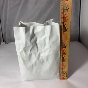 Puede incluir: Un florero de cer&aacute;mica blanca con forma de bolsa de papel arrugada, colocado junto a una regla de madera que muestra que el objeto mide aproximadamente 21,5 cm de altura. El florero tiene un acabado mate y texturas irregulares y plegadas.